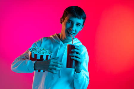 One young boy, student, teen in casual clothes holding huge bucket of popcorn isolated on magenta backgroud in neon light.の写真素材