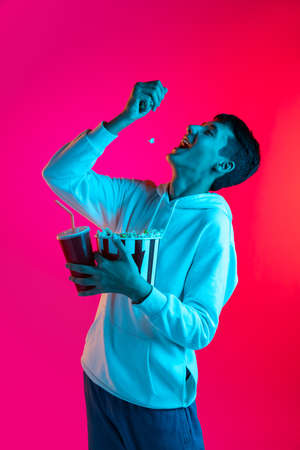 One young boy, student, teen in casual clothes holding huge bucket of popcorn isolated on magenta backgroud in neon light.の写真素材