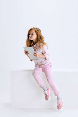 Curiosity. Portrait of little cute girl in casual clothes sitting on big box and using tablet isolated on white background. Childhood, education, study, news, emotions, facial expression conceptの写真素材