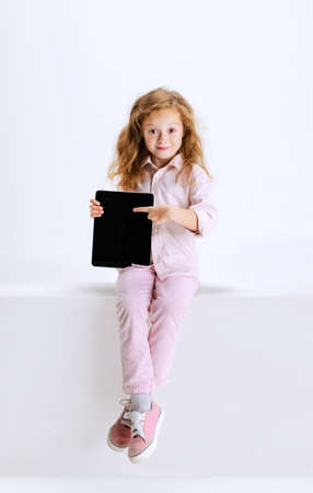 Portrait of little cute girl in casual clothes sitting on big box and using tablet isolated on white studio background.の写真素材