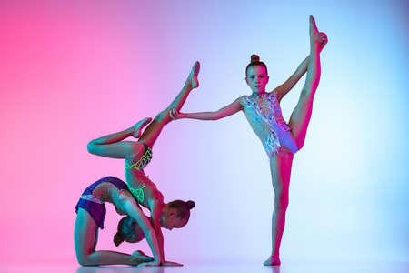 Three little beautiful rhythmic gymnastics artists, girls in sport event isolated on gradient pink blue studio background in neon lightの写真素材