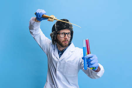 Humorous portrait of young man in image of chemist, doctor wearing white gown and protective gloves isolated on blue background.の写真素材