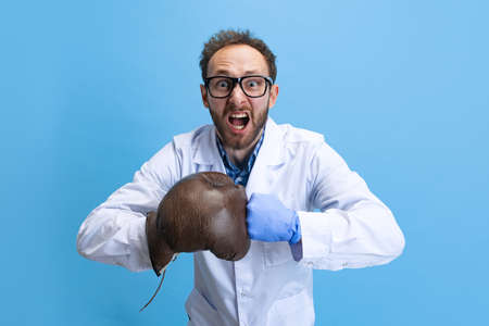 Humorous portrait of young man in image of chemist, doctor wearing white gown and protective gloves isolated on blue background.の写真素材