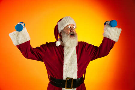 Portrait of happy smiling Santa Claus doing exercises with hand weights isolated on red yellow background in neonの写真素材