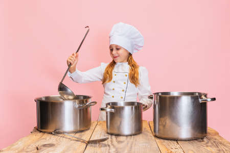 Young little girl in white cook uniform and huge chefs hat at kids kitchen with big pans isolated on pink studio background.の写真素材