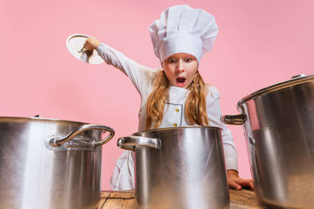Close-up cute little girl in white cook uniform and huge chefs hat at kids kitchen with big pans isolated on pink studio background.の写真素材