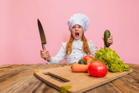 Young little girl in white cook uniform and huge chefs hat cutting fresh vegetables isolated on pink studio background.の写真素材