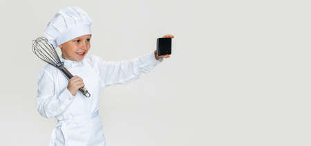 Close-up portrait of little happy boy in white cook uniform and huge chefs hat using phone isolated...