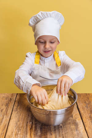 Cute smiling boy in white cook, chef jacket and hat kneads dough isolated on yellow studio background.の写真素材