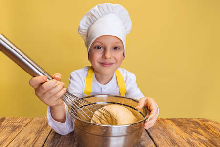 Cute smiling boy in white cook, chef jacket and hat kneads dough isolated on yellow studio background.の写真素材