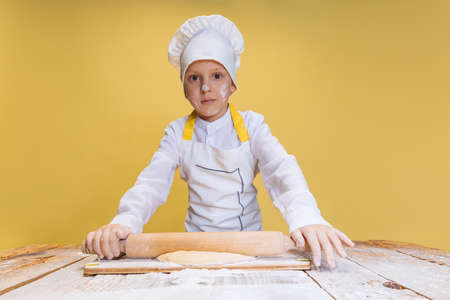One cute boy wearing in white cook, chef jacket and hat kneads dough isolated on yellow studio...