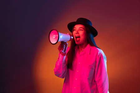 Emotional young girl shouting at megaphone isolated on dark studio background in neon light.