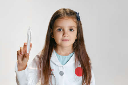 Portrait of cute beautiful girl, child in image of doctor wearing white lab coat with toy syringe isolated on white studio backgroundの写真素材