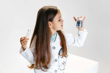 Flyer with cute little girl, child in image of doctor wearing white medicine coat holding pills isolated on white studio backgroundの写真素材