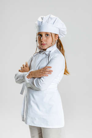 Half-length portrait of little school girl in white cook uniform and huge chefs hat standing with arms crossed isolated on white studio background.の写真素材