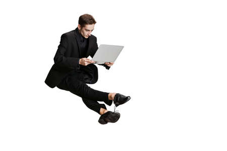 One handsome man in black business suit flying and using laptop isolated on white background. Art, inspiration concept.の写真素材