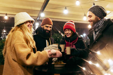 Happy smiling friends with cups of mulled wine having fun, spending time together at winter fair at evening. Holidays, Christmas conceptの写真素材