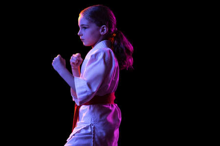 One little girl, young karate in fighting stance isolated over dark background. Concept of sport,...