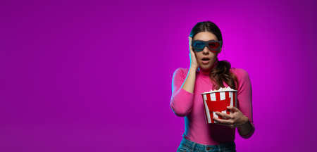 One young beautiful emotional girl watching movie isolated on purple studio background in pink neon...