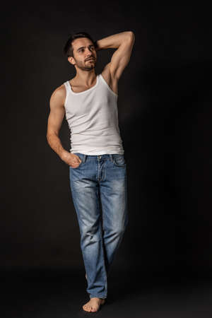 Full-length portrait of young muscular man in white undershirt and jeans posing isolated on dark background.の写真素材