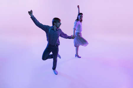 Studio shot of young man and woman in vintage retro style outfits dancing lindy hop isolated on lilac color background in neon lightの写真素材