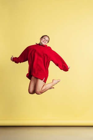 Full-length portrait of young joyful girl in warm red sweater jumping isolated over yellow studio...