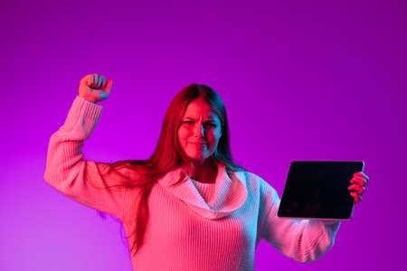 One young happy girl in warm knitted sweater using tablet, gadget isolated over purple studio background in neon light.の写真素材