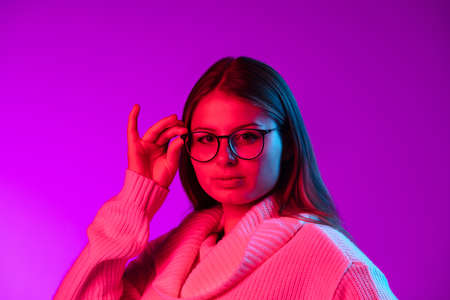Portrait of young pretty girl in warm knitted sweater putting eyeglasses on isolated over purple studio background in neon light.の写真素材