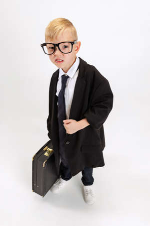 Close-up of cute little boy, kid businessman in huge fathers suit holding big vintage attache case isolated on white studio background.の写真素材