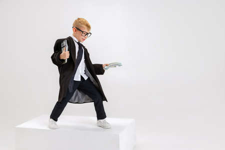 Portrait of emotional little boy, child businessman in huge jacket suit standing on big box isolated on white studio background.の写真素材