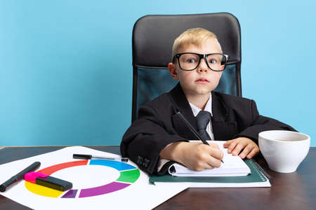 Portrait of little boy, child businessman in huge jacket suit sitting at office table isolated on...