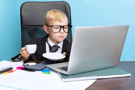 Portrait of little boy, child businessman in huge jacket suit sitting at office table and using laptop isolated on blue studio background.の写真素材