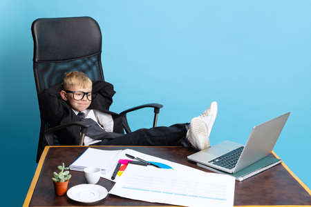 Portrait of little boy, child businessman in huge jacket suit sitting at office table isolated on...