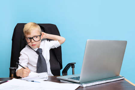 Portrait of little boy, child businessman in eyeglasses and white shirt sitting at office table isolated on blue studio background.の写真素材