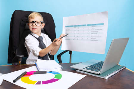 Showing charts. Little boy, child businessman in eyeglasses and white shirt sitting at office table...