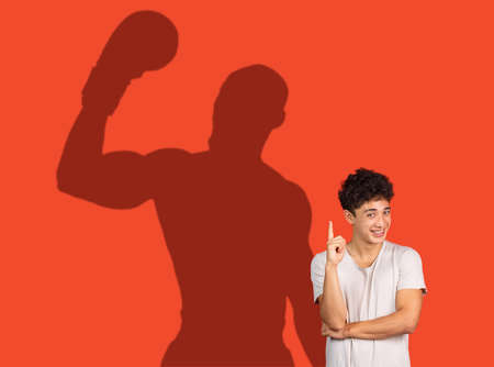 Young man thinking about famous sport future. Conceptual image with smiling boy and shadow of boxer on red wallの写真素材