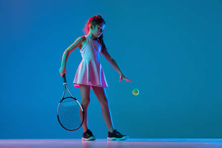One little girl, beginner tennis player posing isolated on blue studio background in neon light....