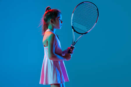 Profile view of little girl, beginner tennis player posing with racket isolated on blue studio background in neon light. Sport, study, childhood conceptの写真素材