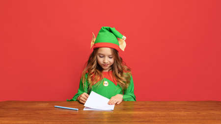Portrait of happy kid dressed like funny gnome or elf sitting at table and writing letter to Santa Claus isolated over red background.の写真素材