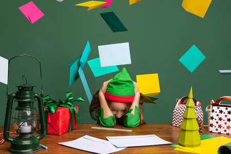 Little happy kid, girl dressed like funny gnome or elf sitting at table and helps Santa Claus to read Christmas mail isolated over green background.の写真素材
