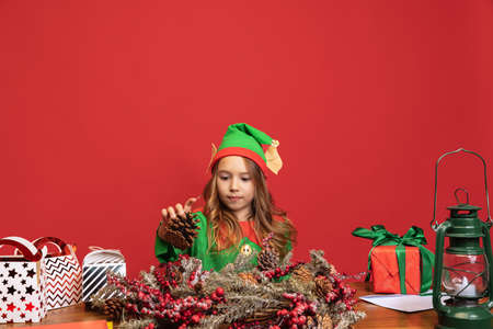 One happy kid dressed like funny gnome or elf sitting at table and making Christmas decorations isolated over red background.の写真素材