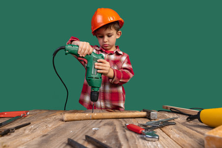 Portrait of cute little boy, kid in image of builder, designer in orange protective helmet using work tools isolated on green backgroundの写真素材