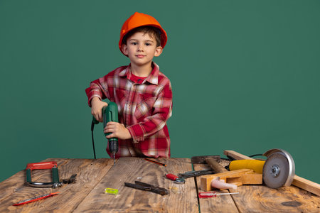 Portrait of cute little boy, kid in image of builder, designer in orange protective helmet using work tools isolated on green backgroundの写真素材