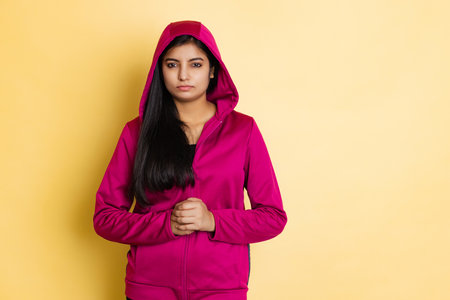 Portrait of young beautiful indian girl, student in magenta color hoody posing isolated on yellow studio backgroundの写真素材