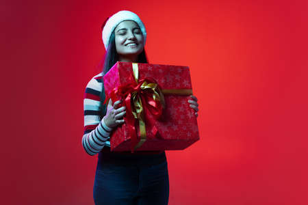 Portrait of young happy girl in warm clothes and Christmas hat holding huge gift box isolated on red...