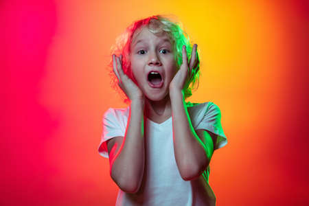 Portrait of little boy, curly surprised kid standing with open mouth isolated on red yellow studio background in neon light, filter.の写真素材