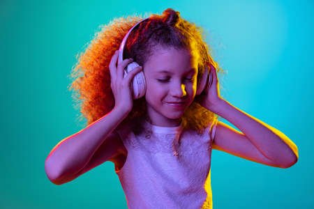 Portrait of little girl, kid listening to music in headphones isolated on blue studio background in neon light, filter.の写真素材