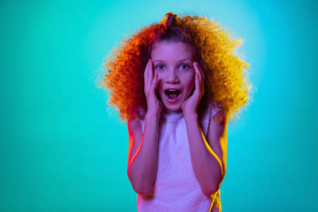 Portrait of little girl, curly surprised kid standing with open mouth isolated on blue studio background in neon light, filter.の写真素材