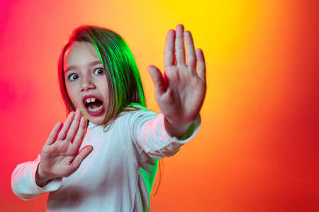 Portrait of little scared girl, kid defends against attack of aggression from outside world isolated on red yellow background in neon lightの写真素材