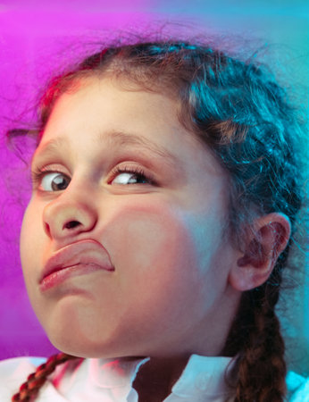 Close-up little girl, cute kid leaning against transparent glass by cheek isolated on blue purple studio background in neon lightの写真素材